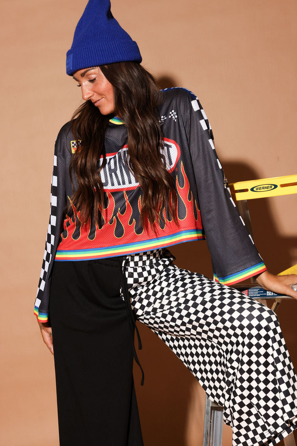 Person wearing a colorful mesh jersey with graphic design, black pants, and a blue beanie against a beige background, leaning on a ladder