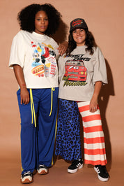 Two people wearing graphic t-shirts and colorful pants against a beige background, frontal view