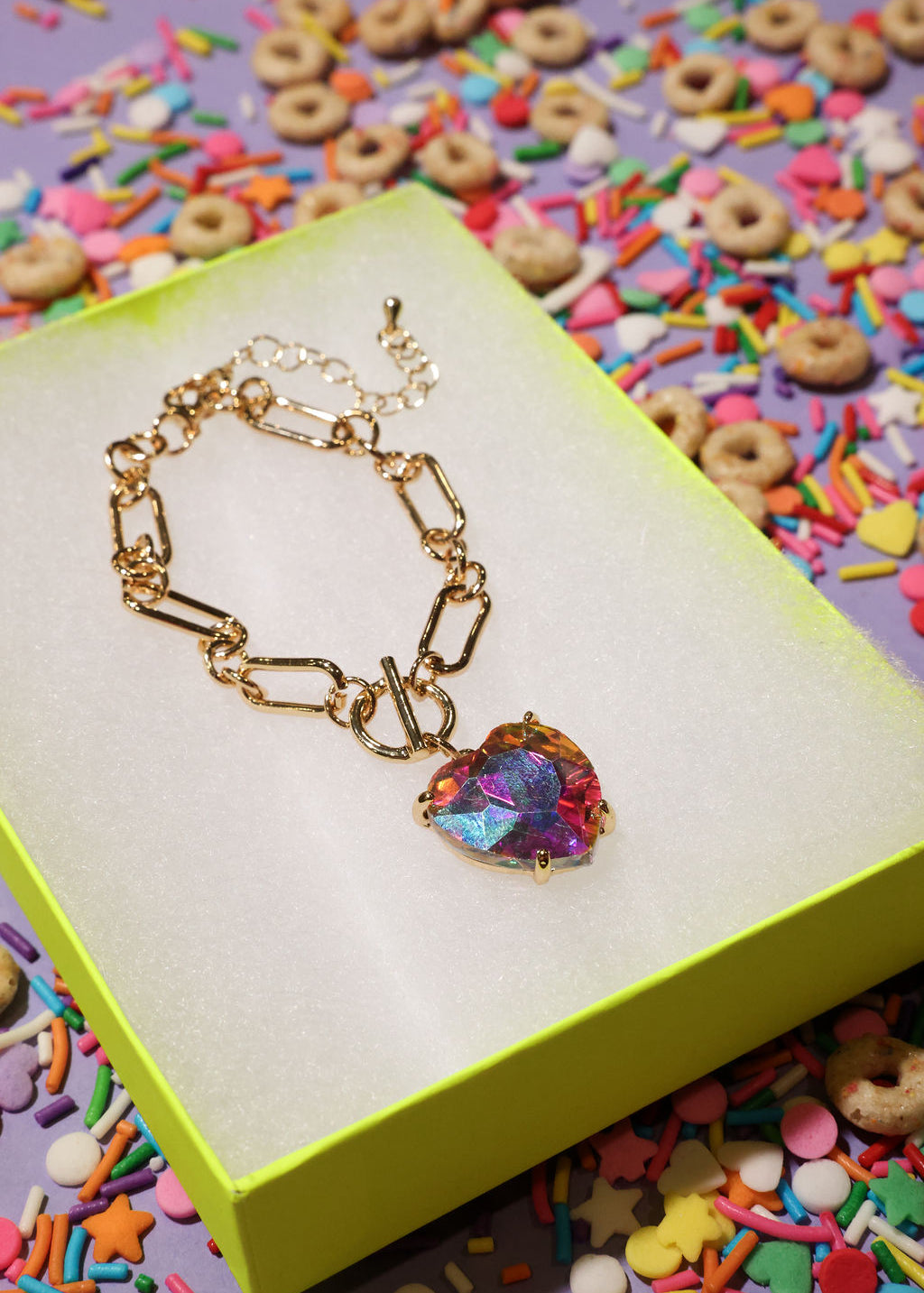 Gold bracelet with a colorful heart charm on a yellow box with a colorful background of sprinkles and cereal