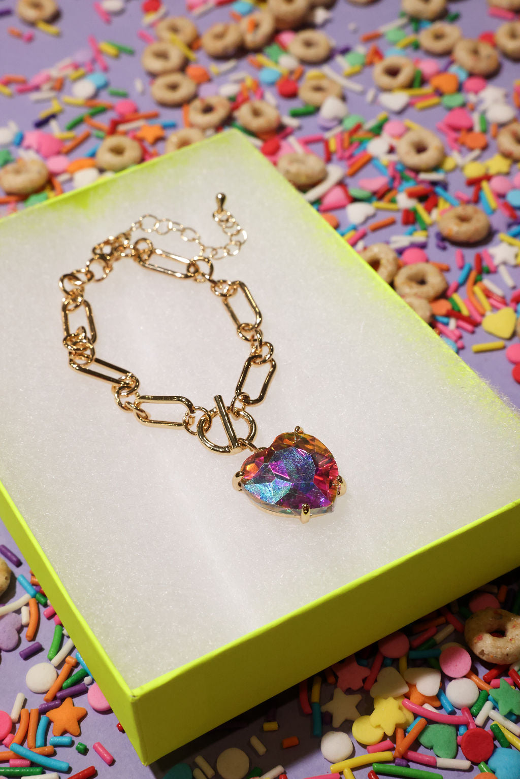 Gold bracelet with a colorful heart charm on a yellow box with a colorful background of sprinkles and cereal