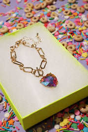 Gold bracelet with a colorful heart charm on a yellow box with a colorful background of sprinkles and cereal