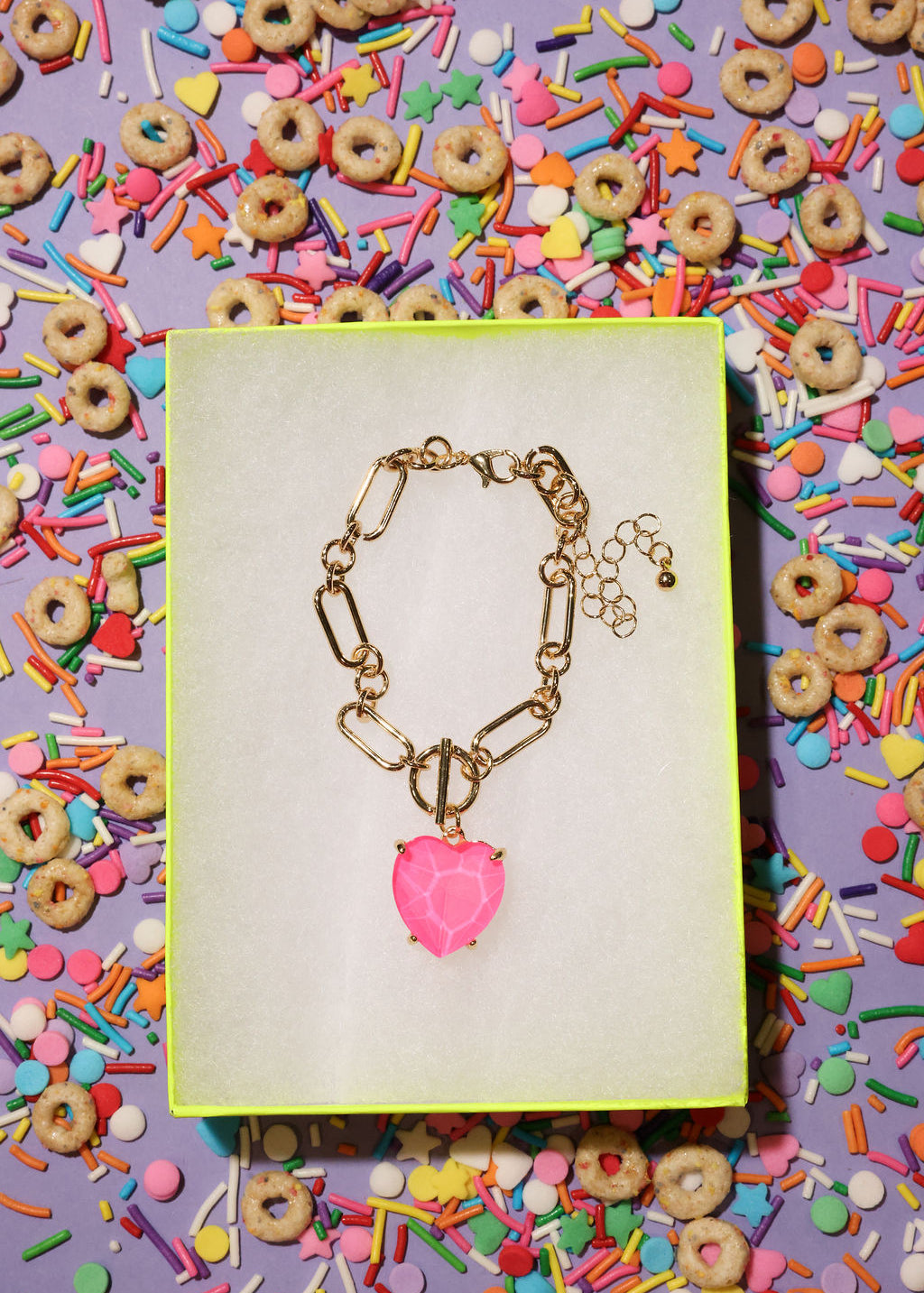 Gold bracelet with a pink heart charm on a yellow card against a colorful background of cereal and sprinkles.