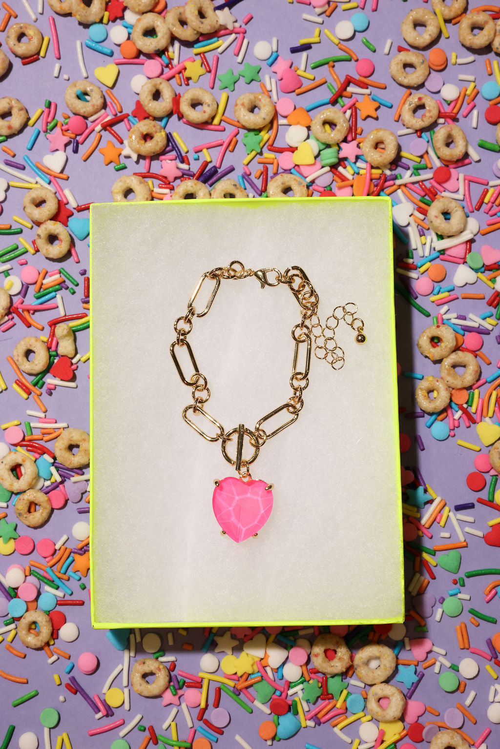 Gold bracelet with a pink heart charm on a yellow card against a colorful background of cereal and sprinkles.