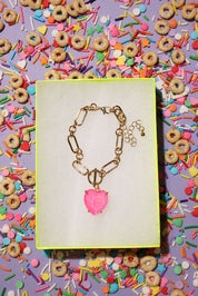 Gold bracelet with a pink heart charm on a yellow card against a colorful background of cereal and sprinkles.