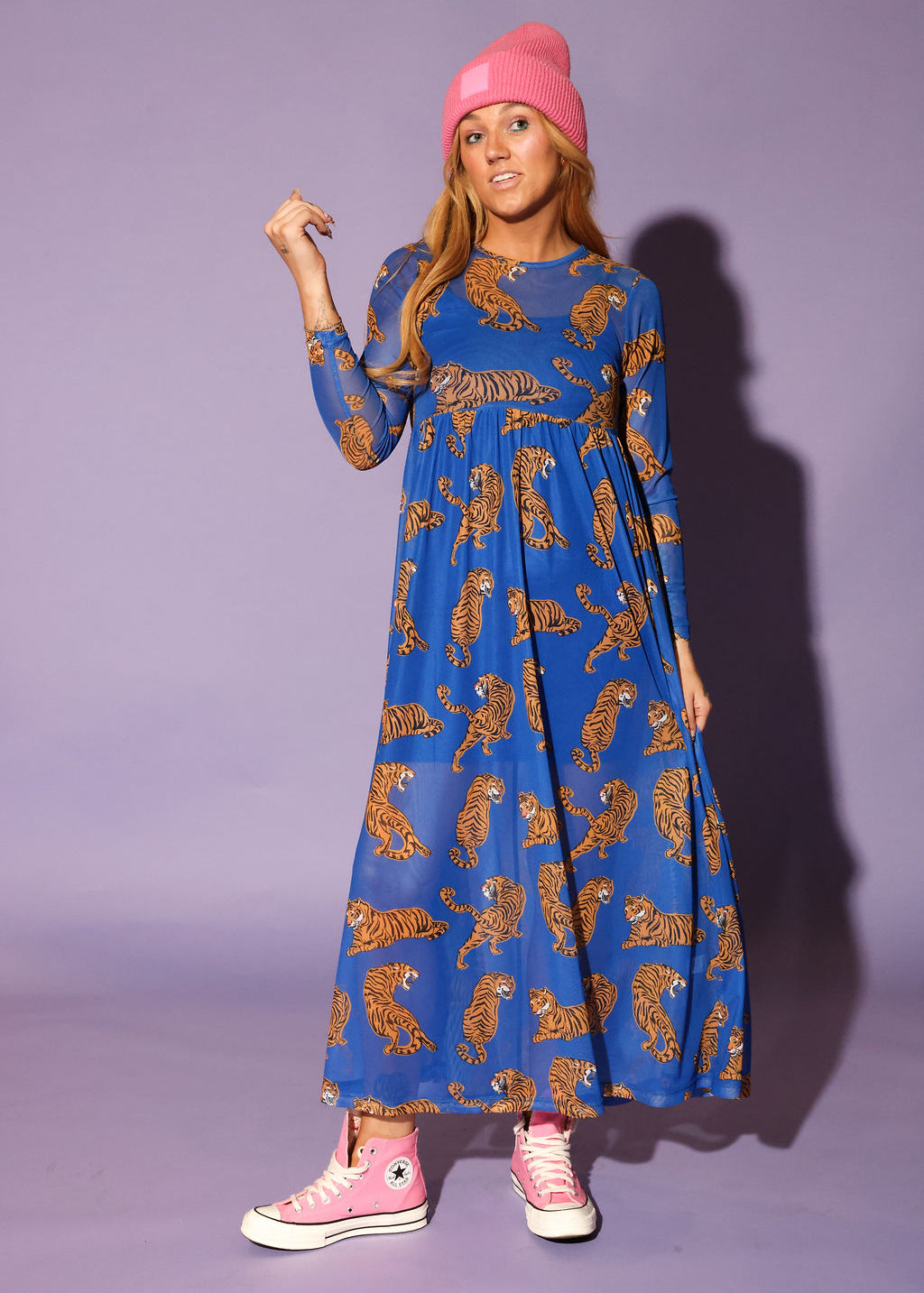 Model wearing a long sleeve blue mesh dress with tiger patterns and a pink beanie, and pink sneakers, against a purple background, frontal view