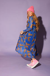 Model wearing a long sleeve blue mesh dress with tiger patterns and a pink beanie, and pink sneakers, against a purple background, side view