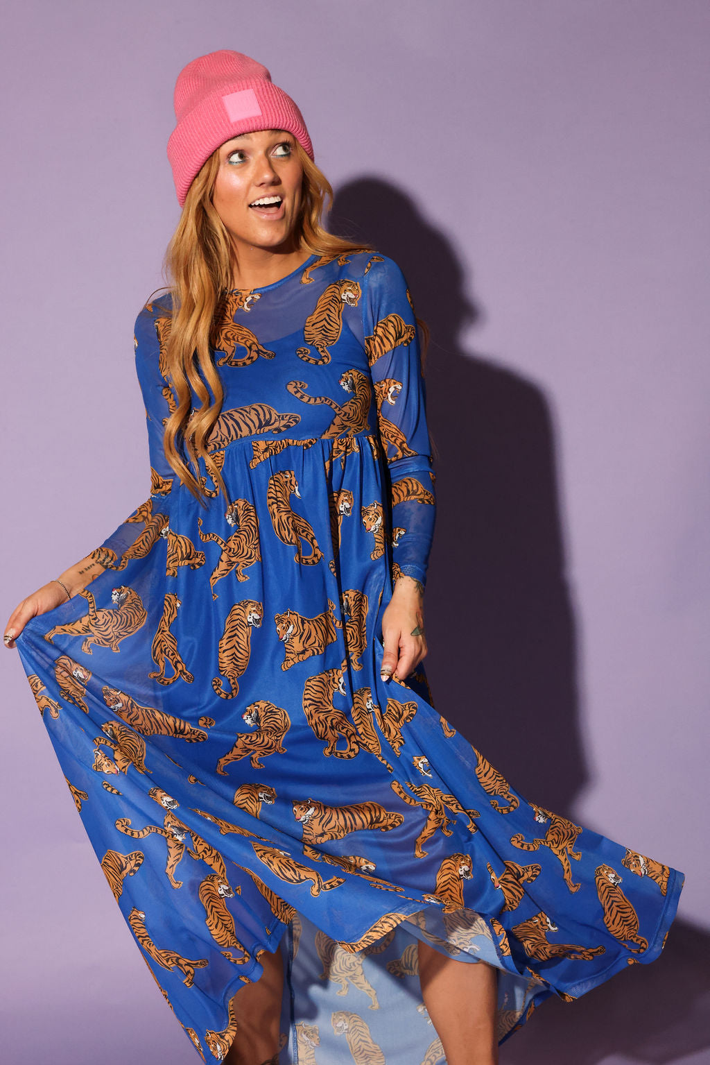 Model wearing a long sleeve blue mesh dress with tiger patterns and a pink beanie, against a purple background, close up frontal view