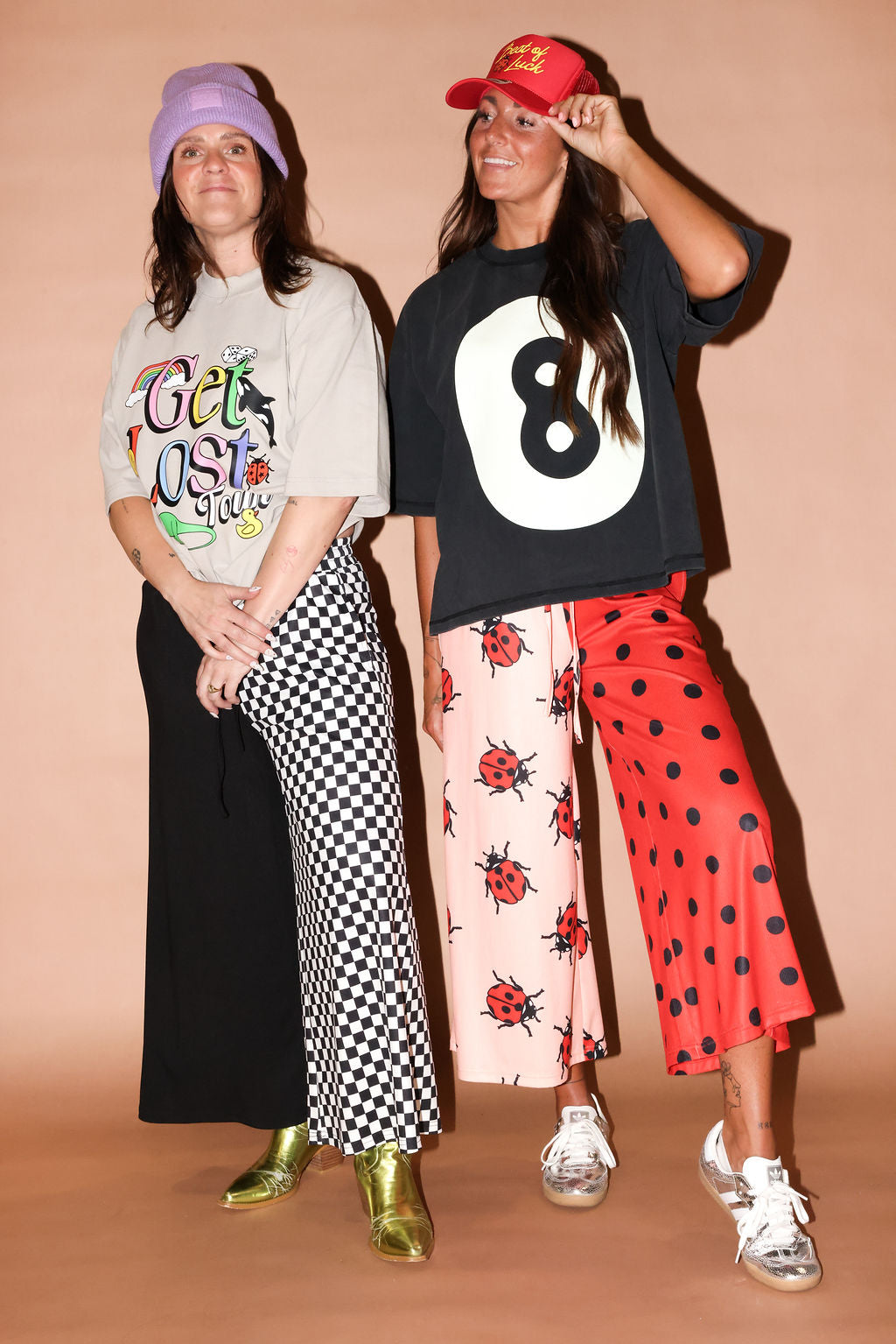 Two women posing in streetwear outfits against a solid peach studio backdrop. The woman on the left wears a lavender beanie, a light gray graphic t-shirt, a black-and-white checkered wide-leg skirt, and metallic gold boots, on a beige background, frontal view