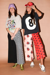 Two women posing in streetwear outfits against a solid peach studio backdrop. The woman on the left wears a lavender beanie, a light gray graphic t-shirt, a black-and-white checkered wide-leg skirt, and metallic gold boots, on a beige background, frontal view