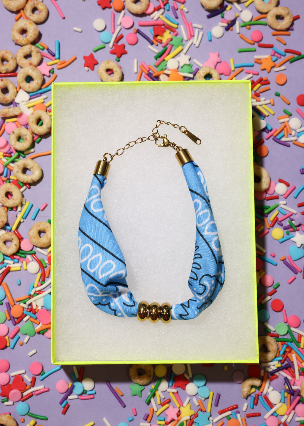 Blue bandana bracelet with gold accents in a box surrounded by colorful cereal and sprinkles.