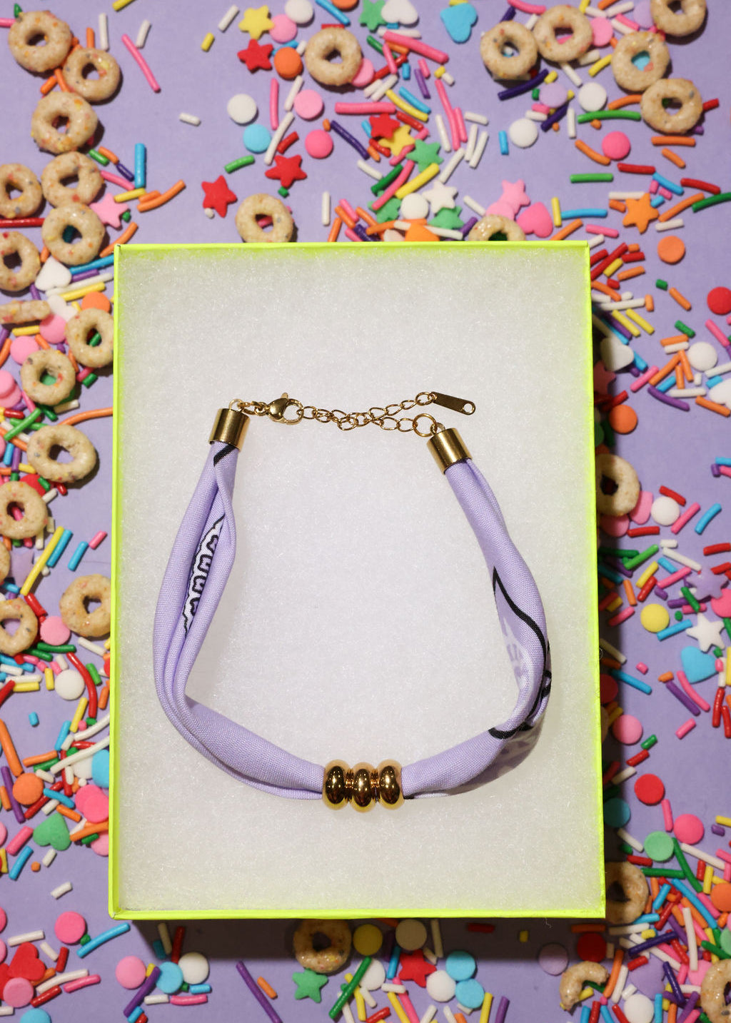 bracelet with a purple bandana cord and gold clasp on a colorful background with donuts and sprinkles