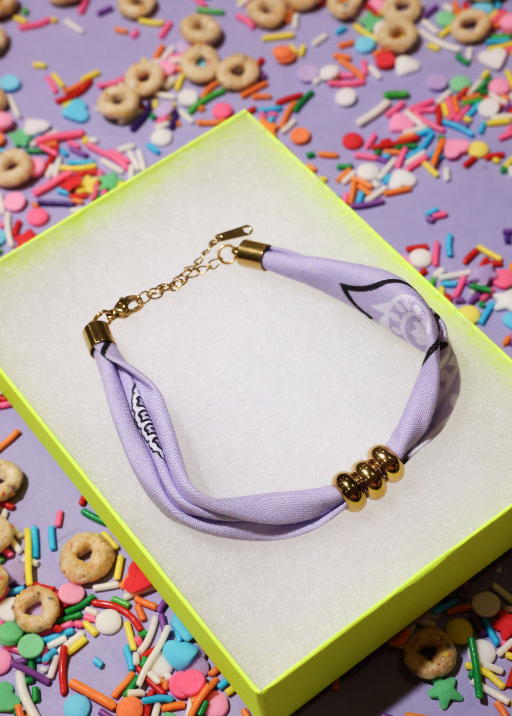 Lavender bracelet with gold accents on a yellow box surrounded by colorful cereal and sprinkles
