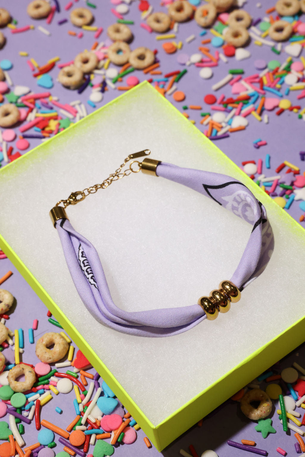 Lavender bracelet with gold accents on a yellow box surrounded by colorful cereal and sprinkles