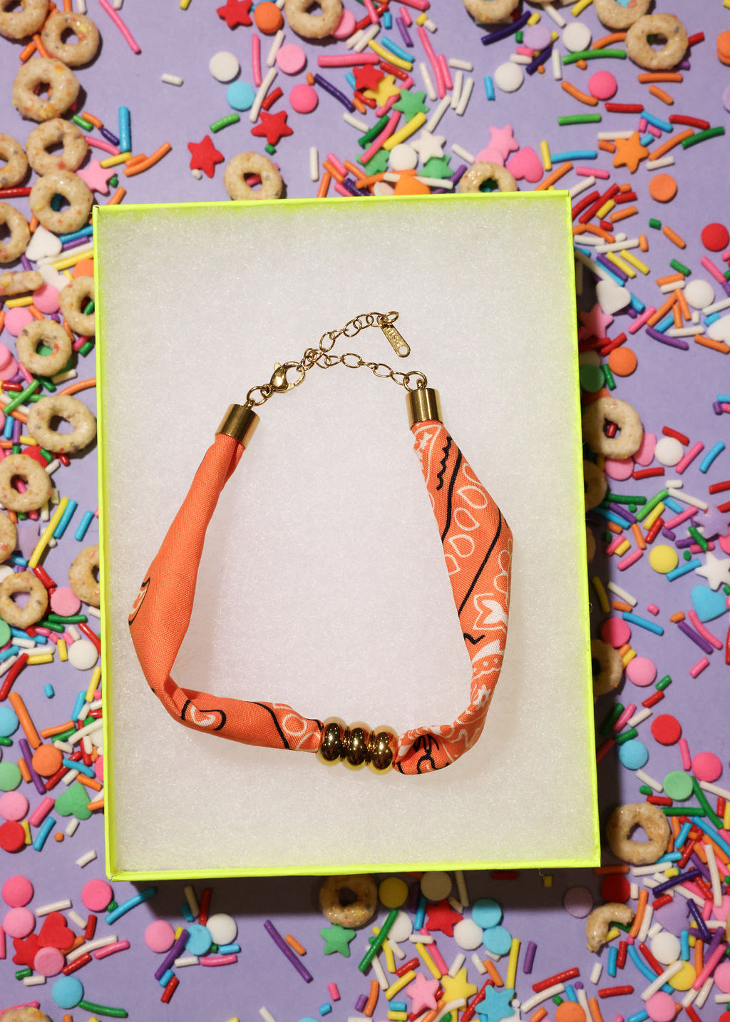 Bracelet with orange and gold accents on a card, surrounded by colorful sprinkles and cereal