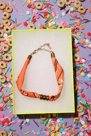 Bracelet with orange and gold accents on a card, surrounded by colorful sprinkles and cereal