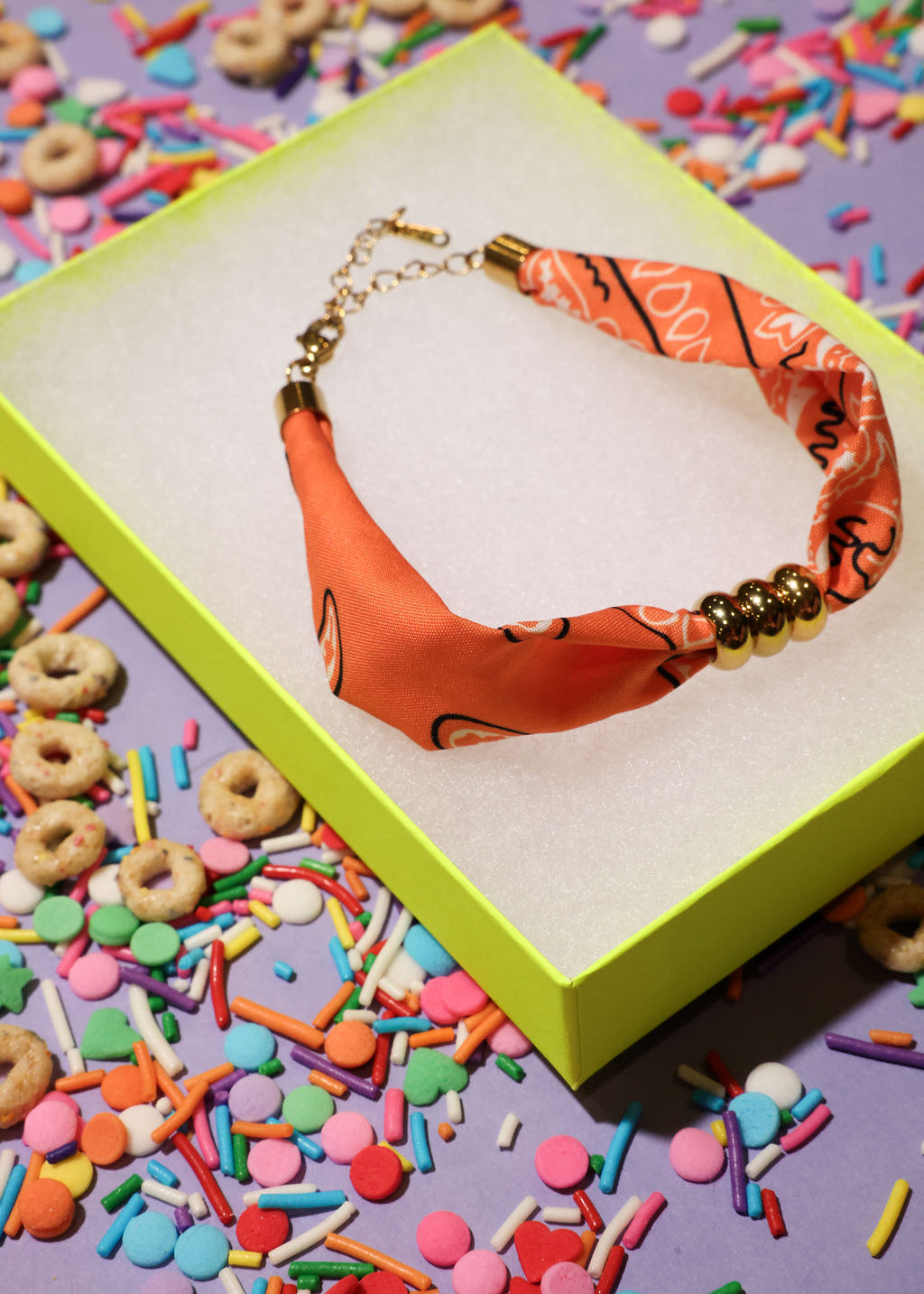 Orange bandana bracelet with gold beads in a green box surrounded by colorful sprinkles and cereal on a purple background