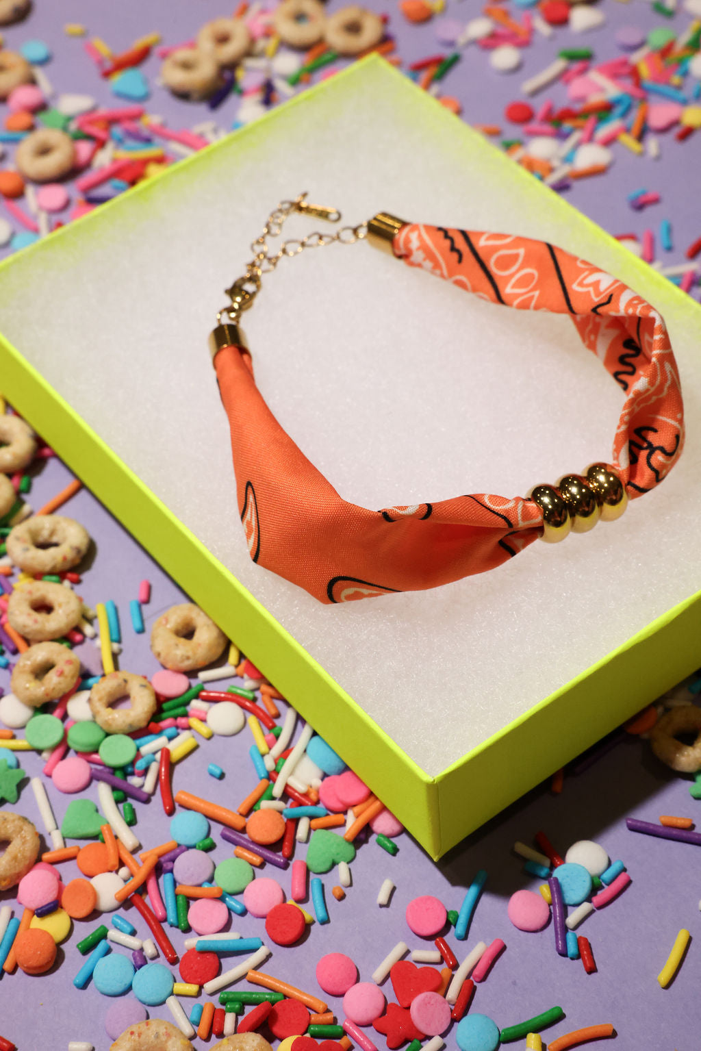 Orange bandana bracelet with gold beads in a green box surrounded by colorful sprinkles and cereal on a purple background