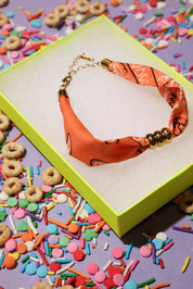 Orange bandana bracelet with gold beads in a green box surrounded by colorful sprinkles and cereal on a purple background