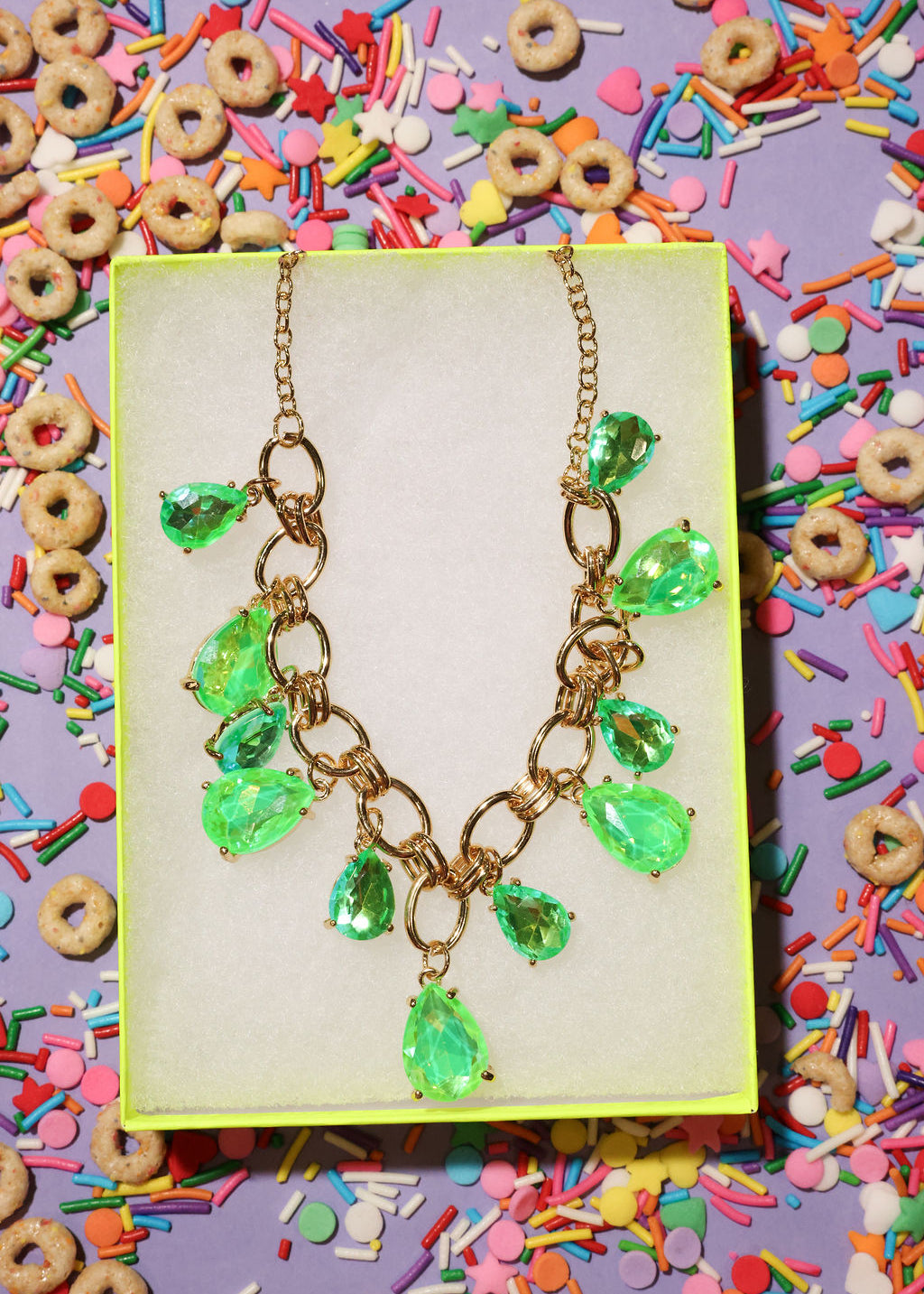 Necklace with green teardrop pendants on a yellow card surrounded by colorful cereal and sprinkles.