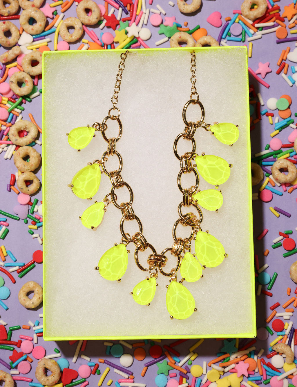 Neon yellow teardrop necklace on a gold chain against a colorful cereal and sprinkles background