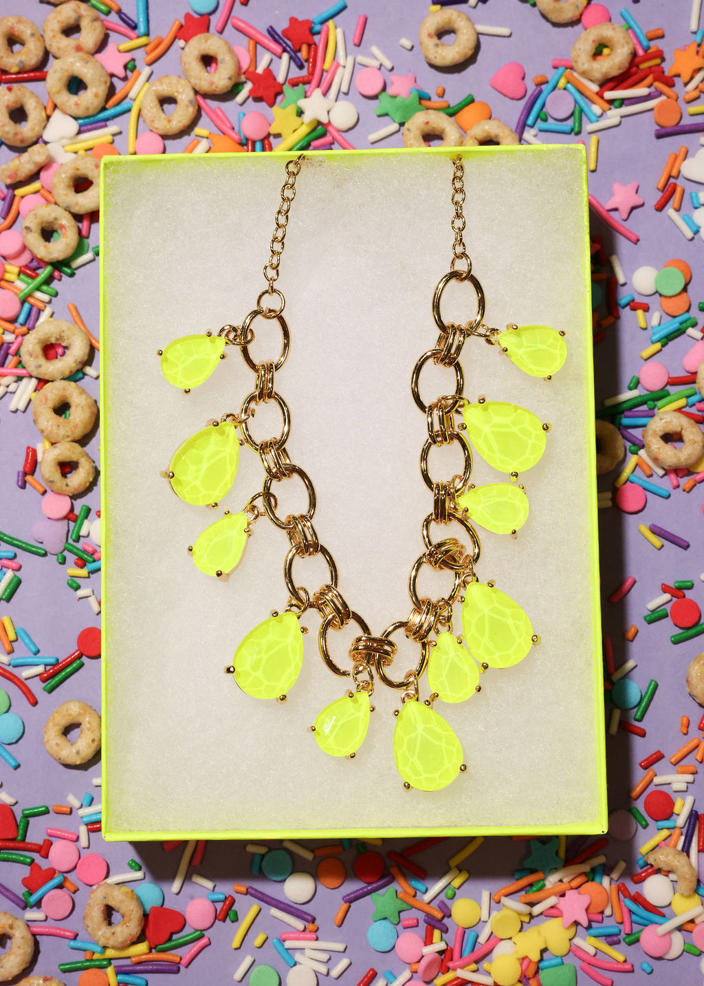 Neon yellow teardrop necklace on a gold chain against a colorful cereal and sprinkles background