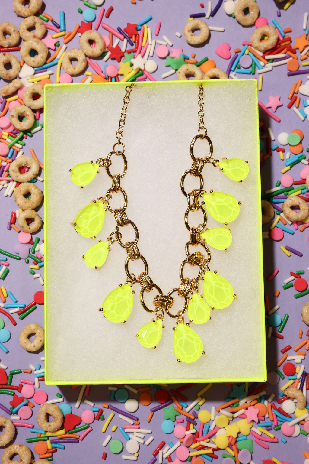 Neon yellow teardrop necklace on a gold chain against a colorful cereal and sprinkles background