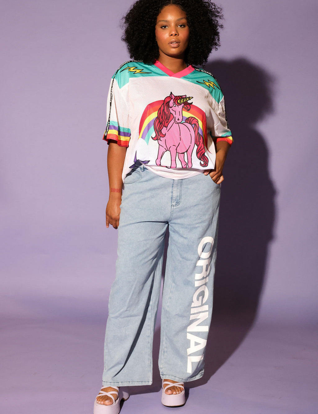 Model wearing a colorful mesh short sleeve jersey with a unicorn and rainbow design, denim pants, on a purple background, frontal view