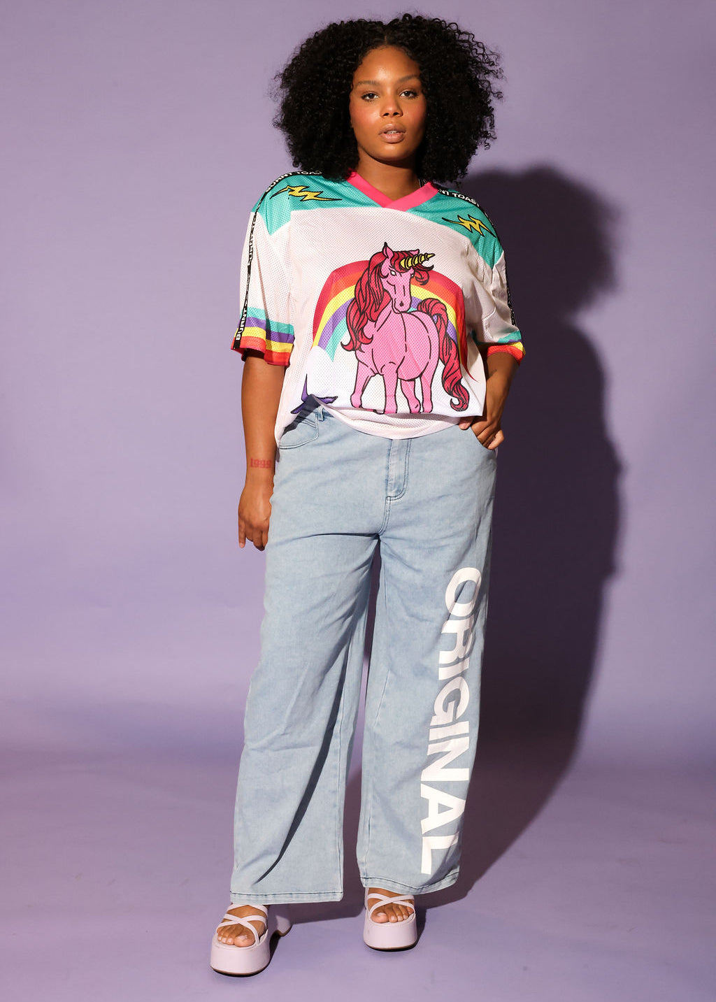 Model wearing a colorful mesh short sleeve jersey with a unicorn and rainbow design, denim pants, on a purple background, frontal view