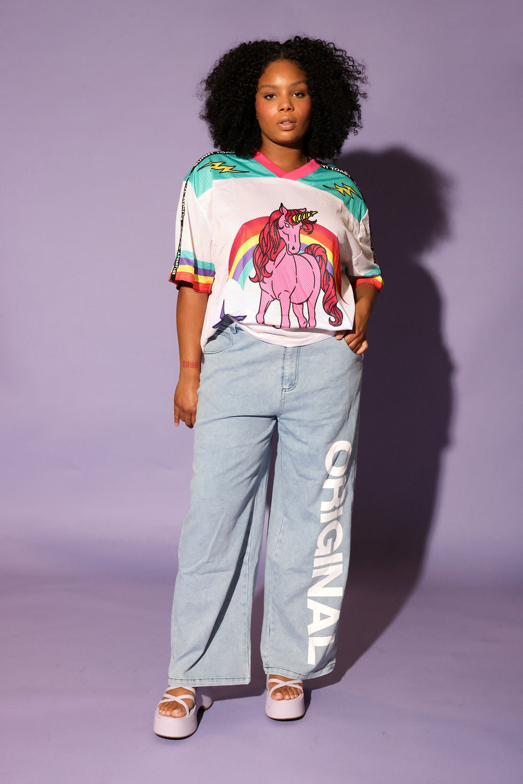 Model wearing a colorful mesh short sleeve jersey with a unicorn and rainbow design, denim pants, on a purple background, frontal view