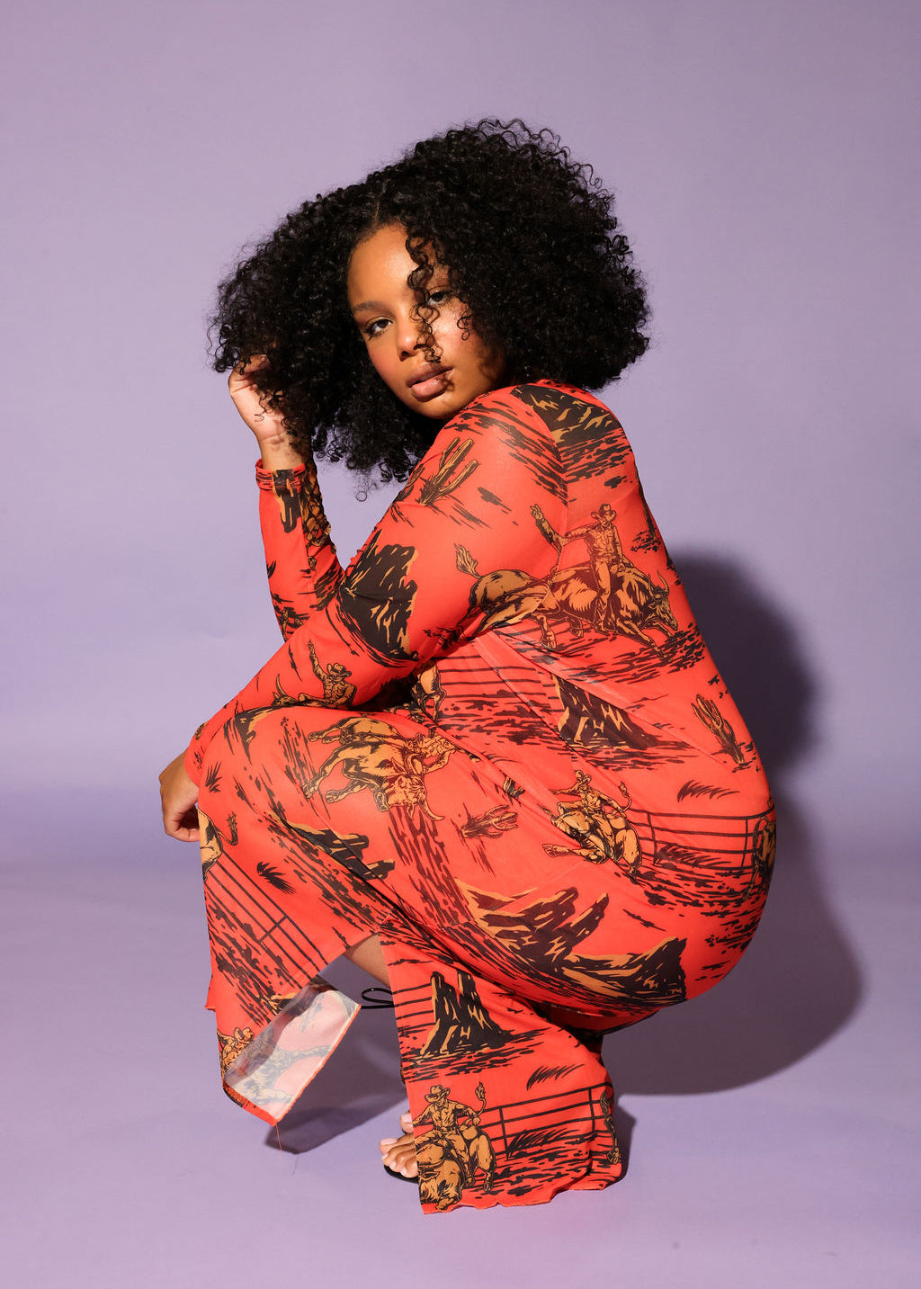 A model with curly black hair crouches in a studio setting against a solid lavender background, wearing a long-sleeve dress in a vibrant orange-red color with a black graphic print, frontal view