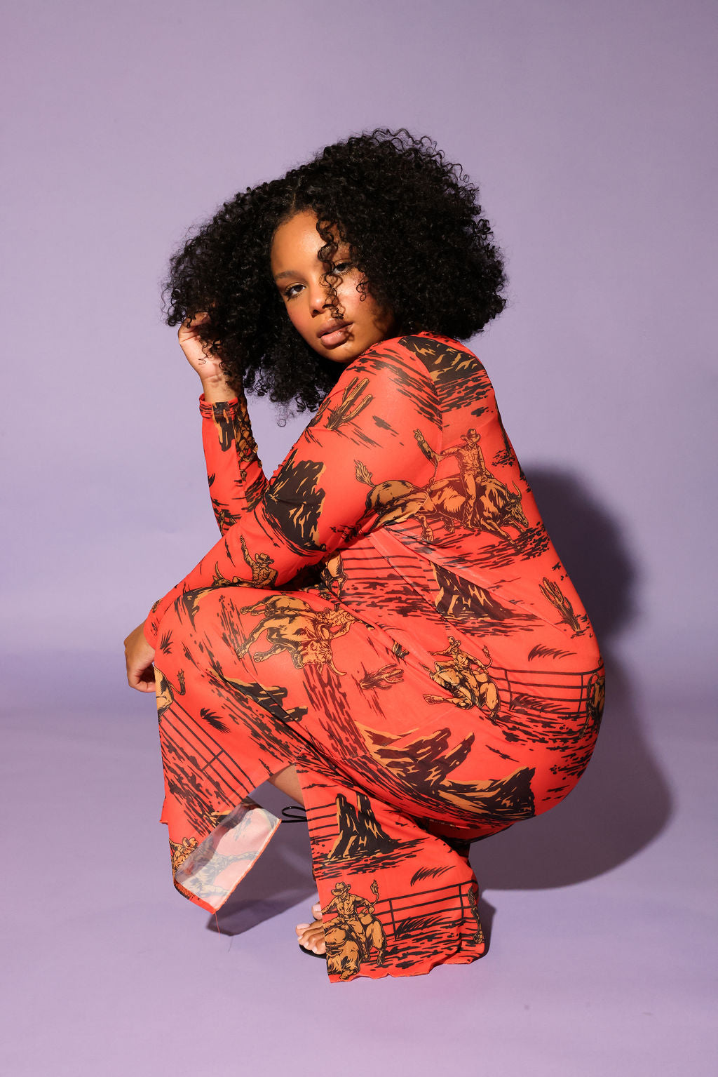 A model with curly black hair crouches in a studio setting against a solid lavender background, wearing a long-sleeve dress in a vibrant orange-red color with a black graphic print, frontal view