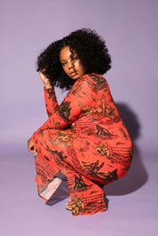 A model with curly black hair crouches in a studio setting against a solid lavender background, wearing a long-sleeve dress in a vibrant orange-red color with a black graphic print, frontal view