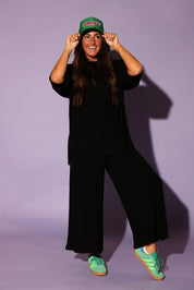 Front view of a model wearing a black ribbed two piece set, including a loose fitting, short sleeve top and high waisted pants. The model is standing with a relaxed posture and wearing green sneakers