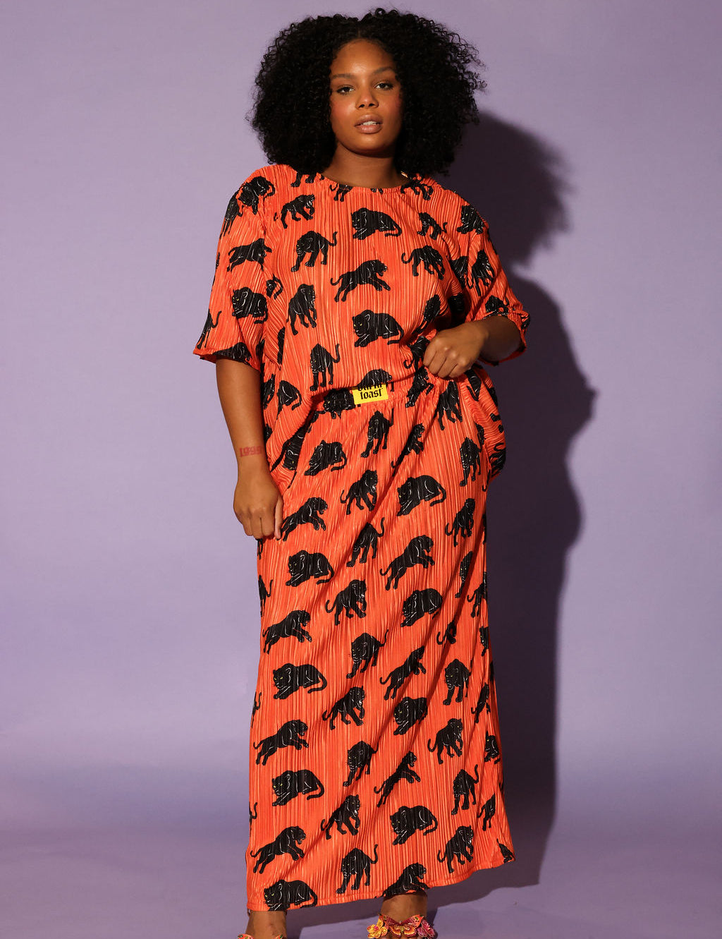 Model wearing an orange plisse short sleeve and skirt set with black animal prints, black beanie, and black and white boots, on a purple background, frontal view