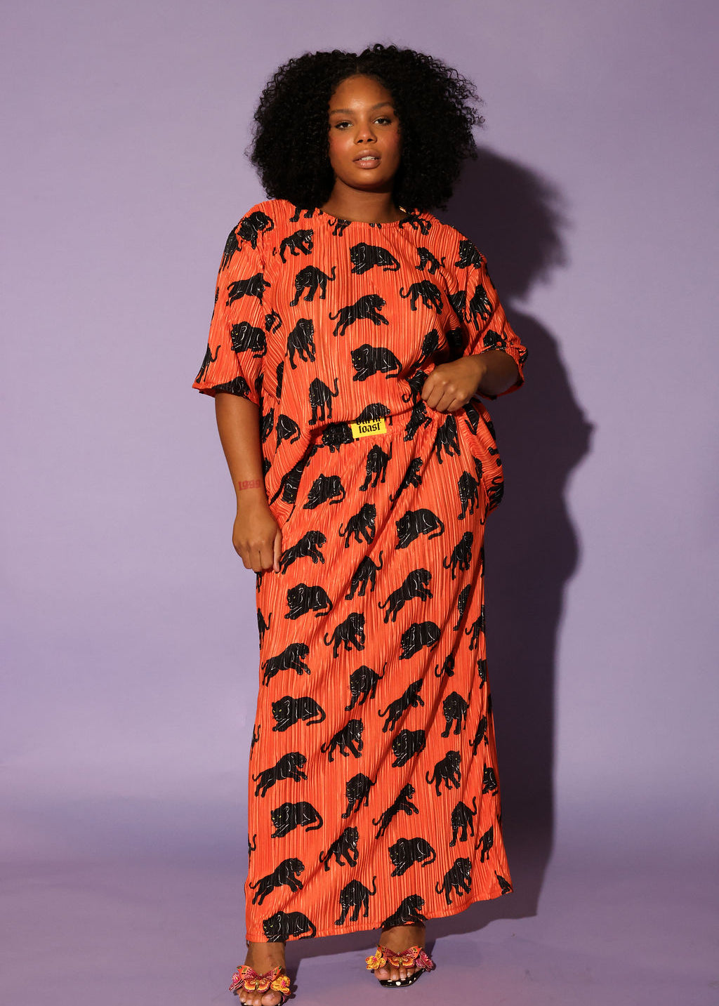 Model wearing an orange plisse short sleeve and skirt set with black animal prints, black beanie, and black and white boots, on a purple background, frontal view