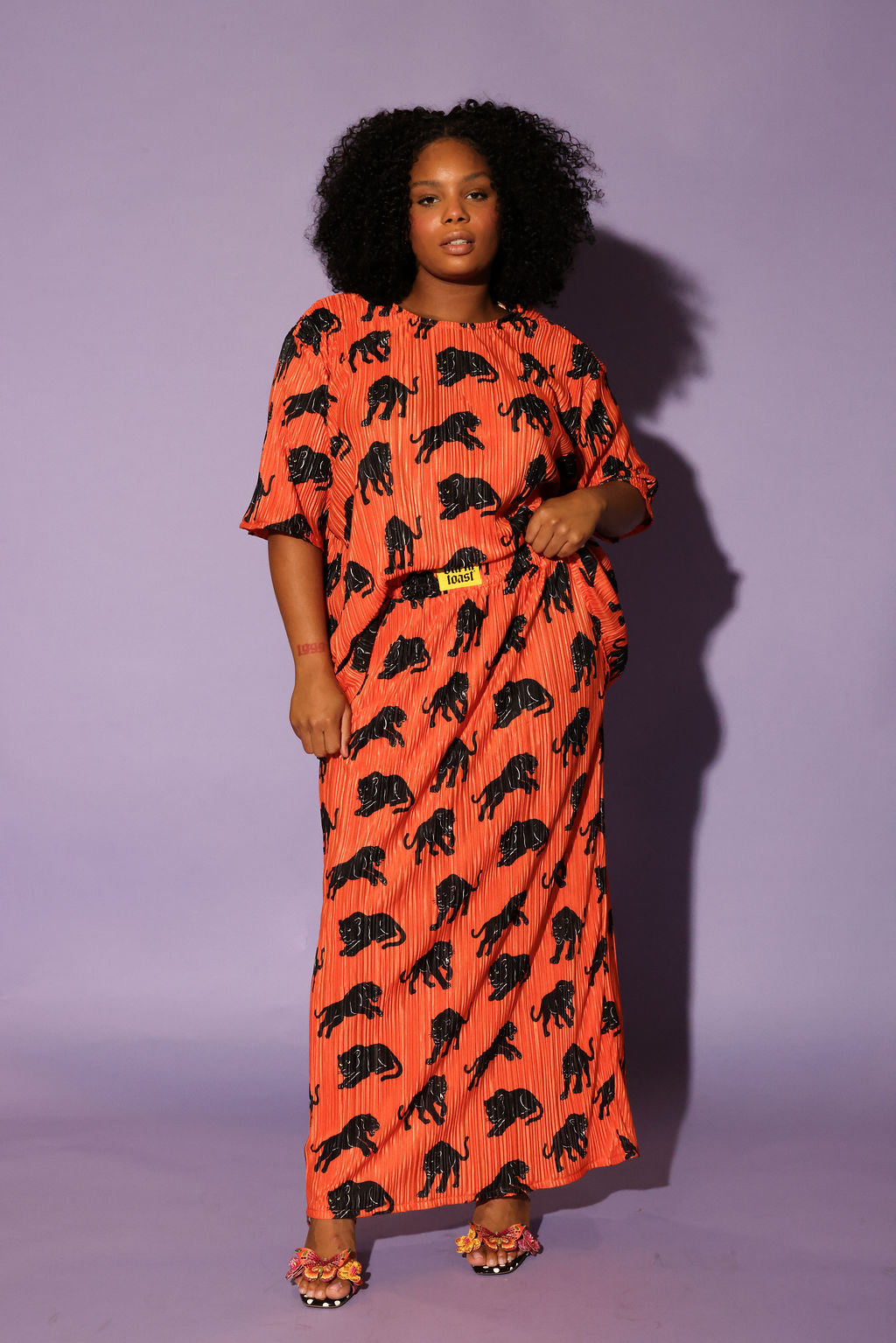 Model wearing an orange plisse short sleeve and skirt set with black animal prints, black beanie, and black and white boots, on a purple background, frontal view