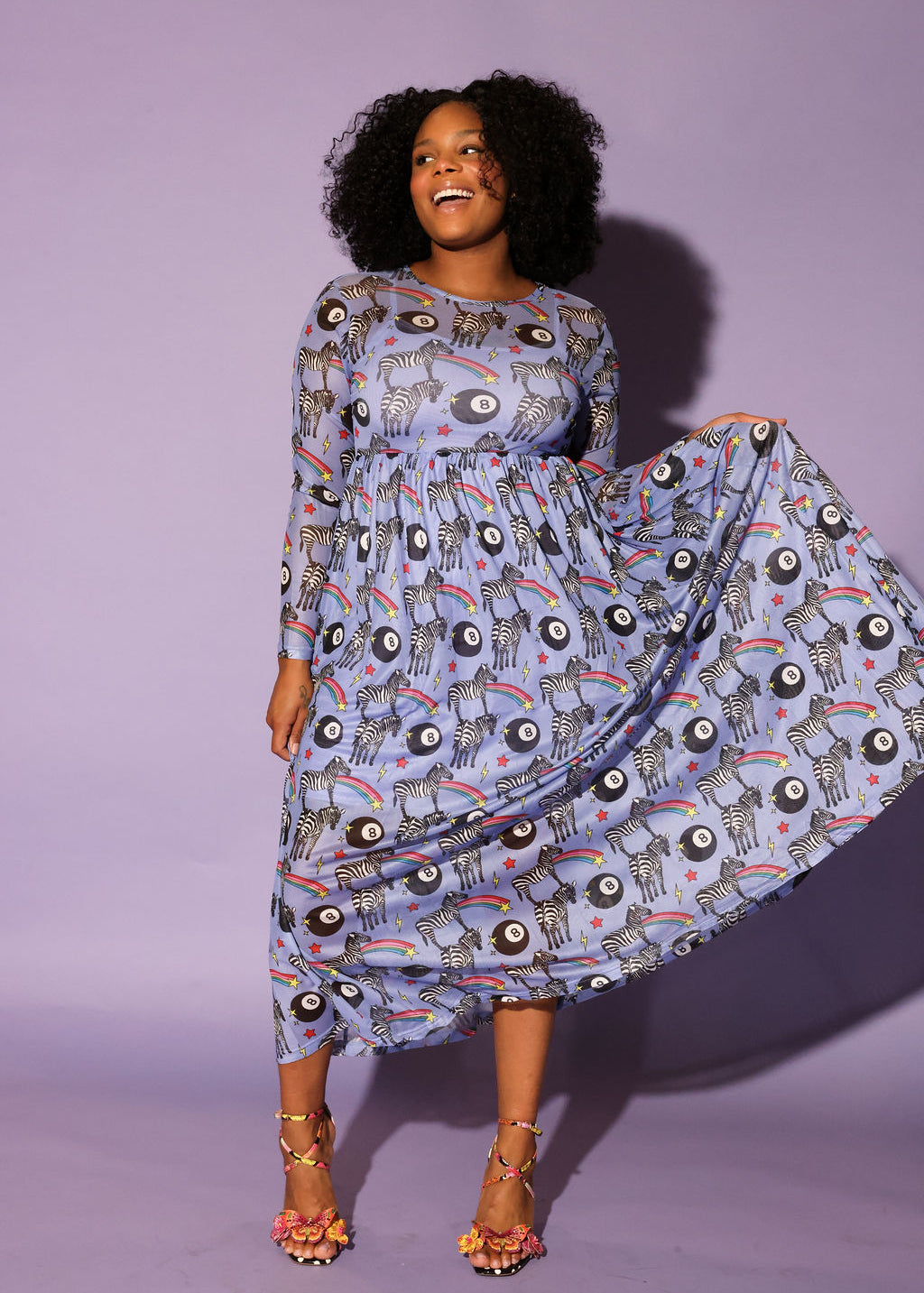 Model wearing a black trucker hat with red and yellow circle logo, long purple mesh dress with a zebra, rainbow and 8 ball design print, in front of a purple background, full frontal view