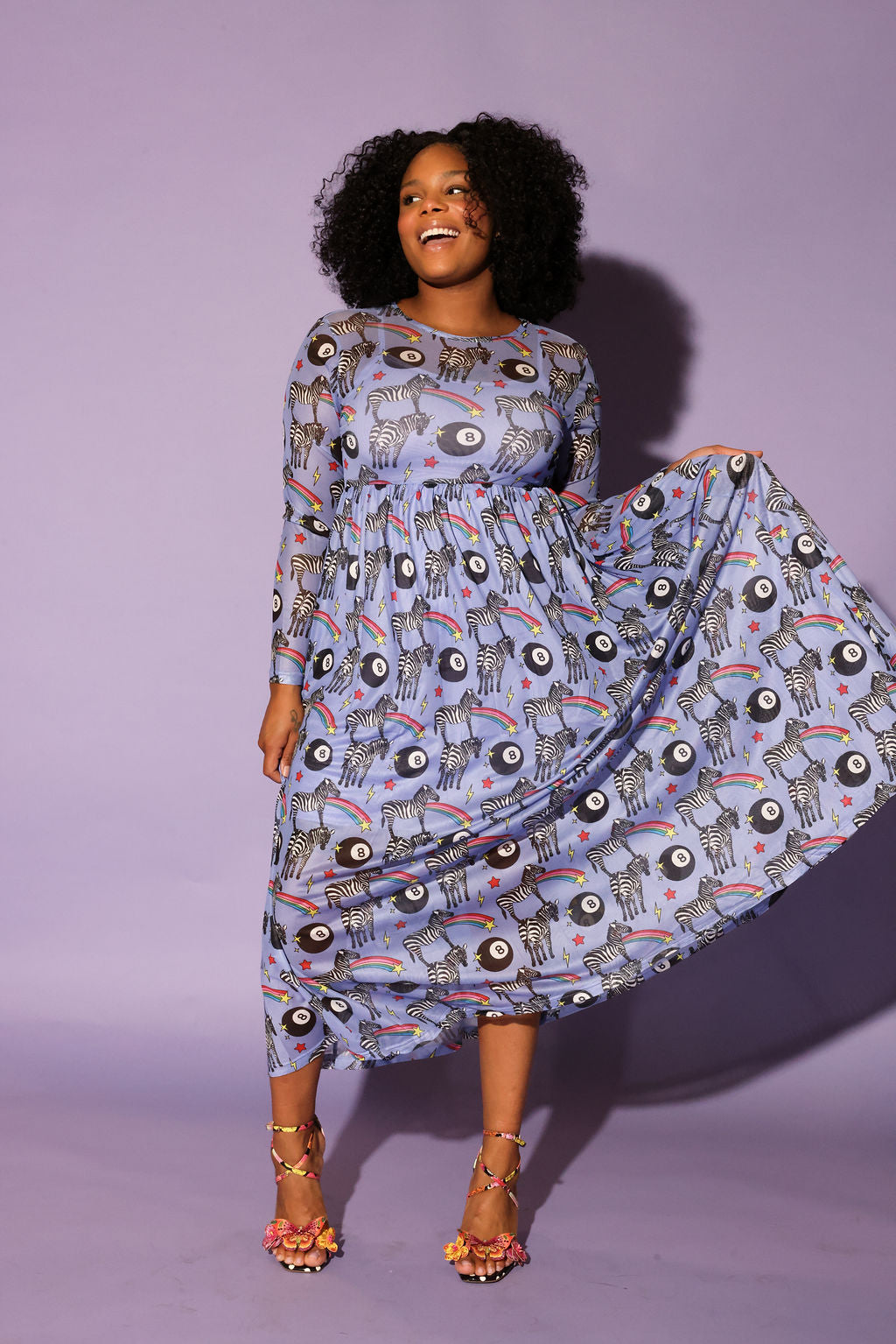Model wearing a black trucker hat with red and yellow circle logo, long purple mesh dress with a zebra, rainbow and 8 ball design print, in front of a purple background, full frontal view