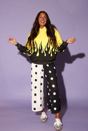 A woman modeling a black and yellow sweatshirt with a flame pattern, paired with wide-leg pants split down the middle with black and white polka dots, against a solid purple background, frontal view