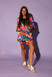 Model wearing a colorful floral jacket with polka dot collar, and matching shorts, a purple background, frontal view