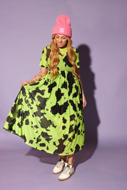 Model wearing a green and black cow print dress with a pink beanie, on a purple background, frontal view