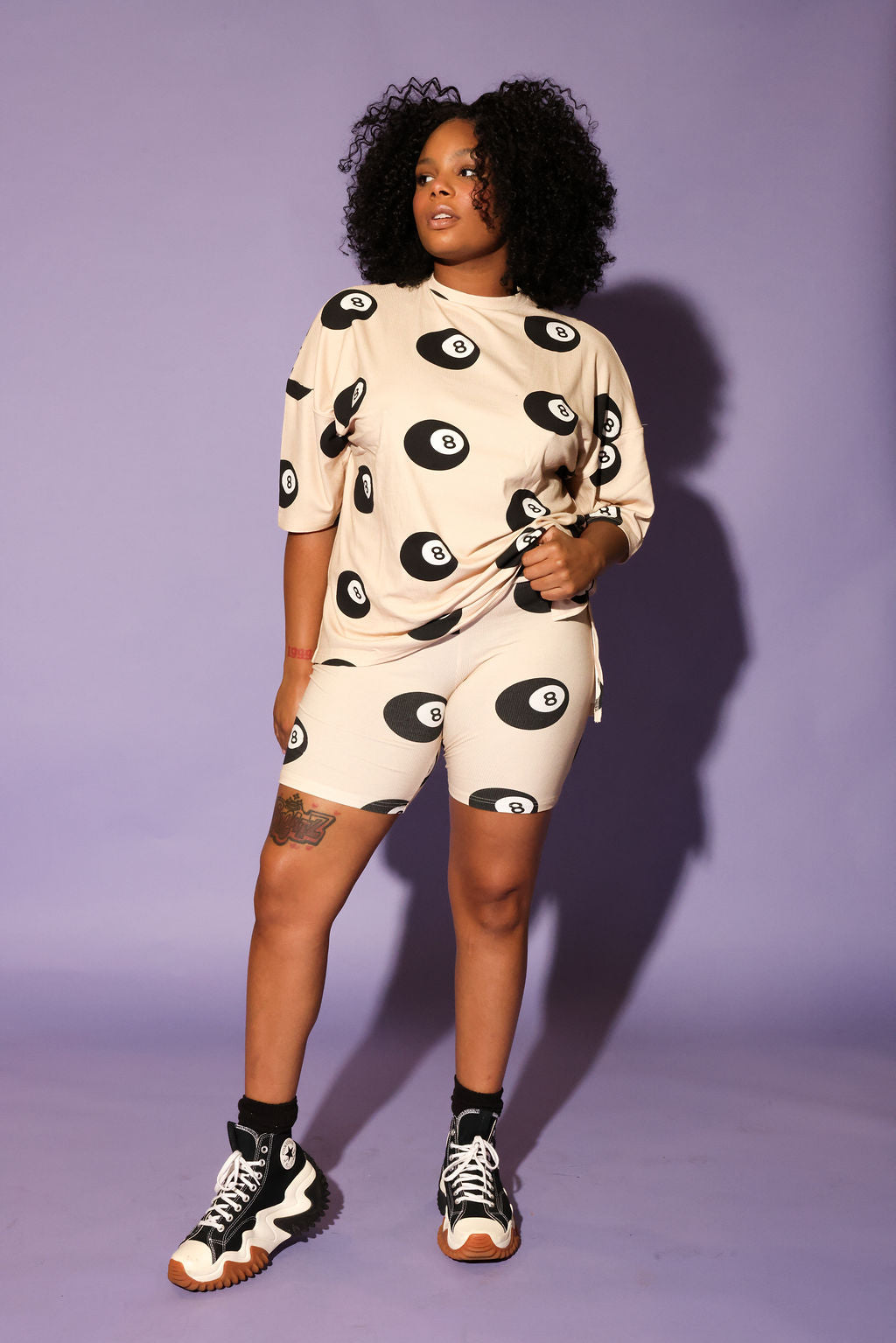 Model wearing a beige biker short, and short sleeve set with black 8 balls throughout, against a purple background, full frontal view
