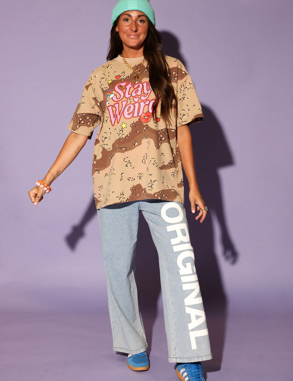 Person wearing a camouflage shirt with 'Stay Weird' text, light blue pants with 'Original' text, and blue sneakers against a purple background, frontal view