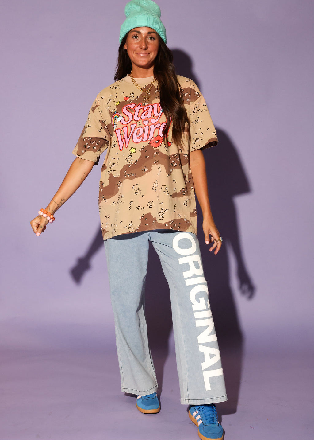 Person wearing a camouflage shirt with 'Stay Weird' text, light blue pants with 'Original' text, and blue sneakers against a purple background, frontal view