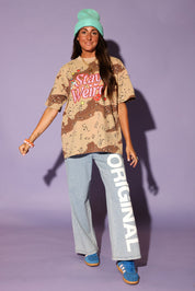 Person wearing a camouflage shirt with 'Stay Weird' text, light blue pants with 'Original' text, and blue sneakers against a purple background, frontal view