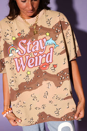 Person wearing a camouflage t-shirt with 'Stay Weird' text and graphics.