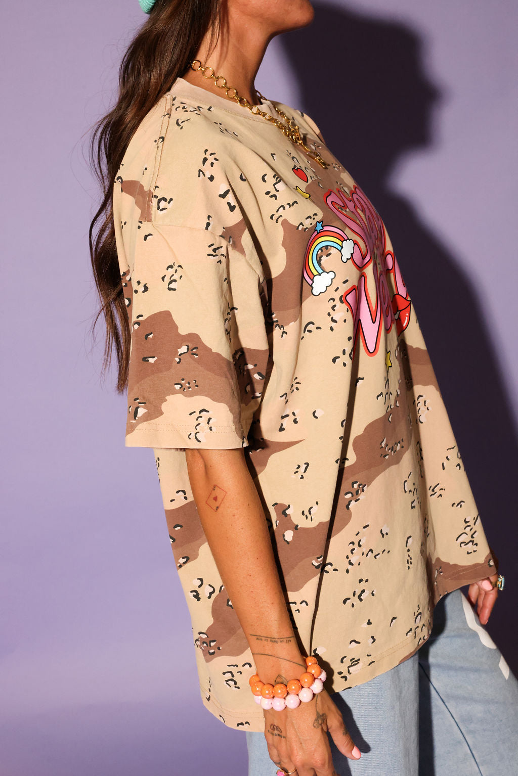 Person wearing a camouflage shirt with 'Stay Weird' text, light blue pants against a purple background, side view