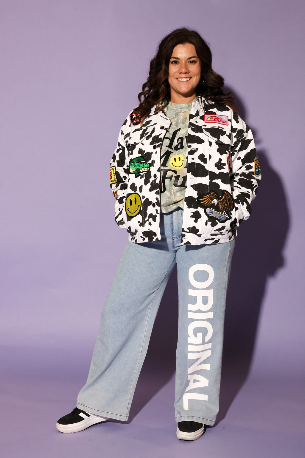 A woman modeling a black and white cow-print patchwork bomber jacket with various graphic patches, worn over a gray t-shirt and paired with wide-leg light blue jeans featuring 'ORIGINAL' printed vertically on the leg, set against a solid lavender studio background, frontal view