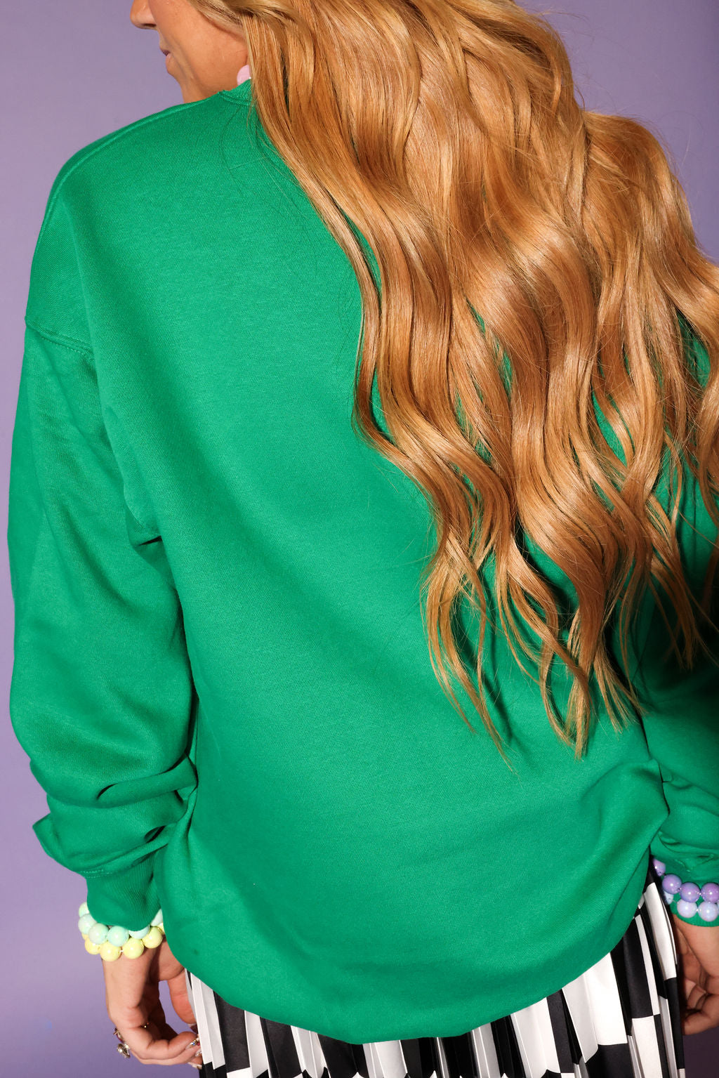 Person wearing a green sweater with long blonde hair, standing against a purple background, back view