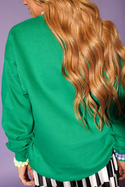 Person wearing a green sweater with long blonde hair, standing against a purple background, back view